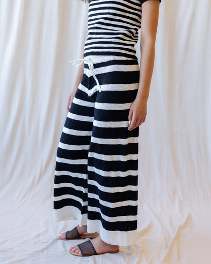 A person wearing a black and white horizontally striped knit dress with a drawstring waist, paired with brown slide sandals, standing against a draped white fabric backdrop.