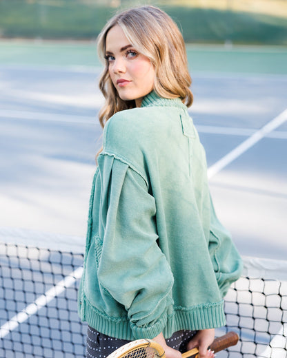 A woman on a tennis court wearing a sage green knit sweater, holding a tennis racket.