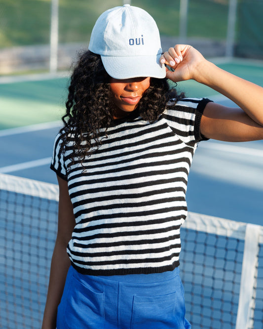 A woman on a tennis court wearing a white baseball cap with the word 'oui' in blue, a black and white horizontally striped knit top, and bright blue skirt or shorts.