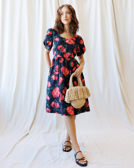 A woman stands against a draped white fabric backdrop wearing a dark navy midi dress with a bold red and pink floral print, puff sleeves, and a gathered waist. She is holding a small woven straw handbag and wearing black strappy flat sandals.