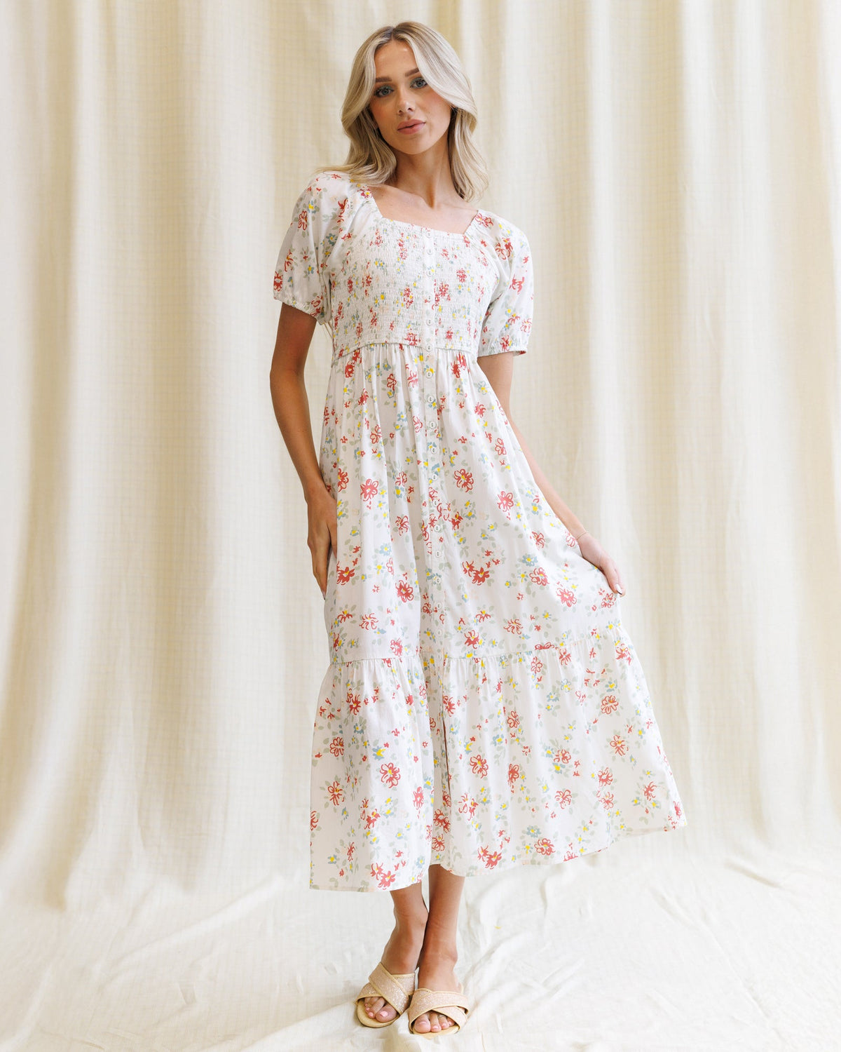 A woman models a white midi dress with a pink and green floral print, featuring puff sleeves, a smocked bodice, and tiered skirt, standing against a draped cream fabric backdrop.