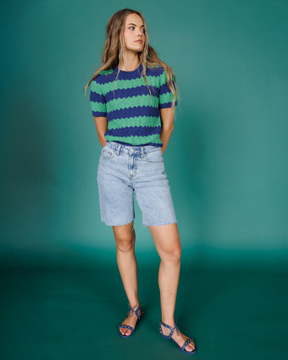 A woman modeling a short-sleeve knit top with horizontal wavy stripes in navy blue and green, paired with light-wash denim shorts and blue strappy sandals, set against a solid teal studio backdrop.