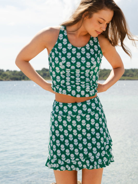 A woman wearing a green two-piece swimsuit with a white floral print stands by the water, looking down. The set includes a halter crop top and a matching ruffled mini skirt.