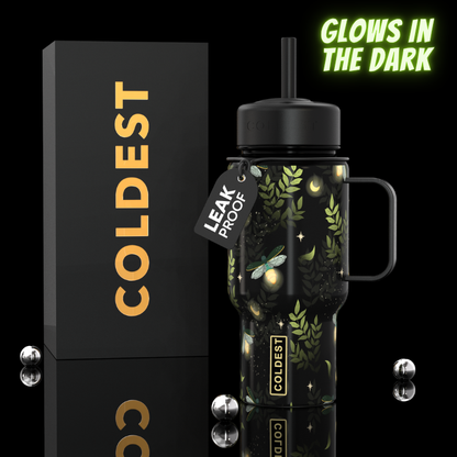 A black handled insulated tumbler with a lid and straw, decorated with glowing green leaves, stars, and moon phases, shown next to its black box with gold 'COLDEST' lettering. A tag reads 'LEAK PROOF' and the image includes a 'GOWS IN THE DARK' badge.