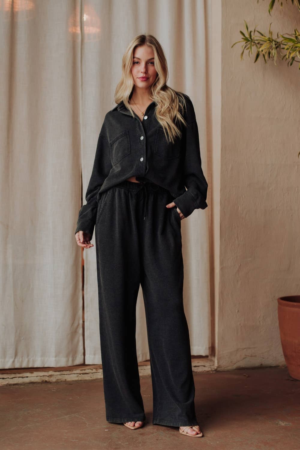 Woman wearing a black outfit standing against a beige wall with a curtain.