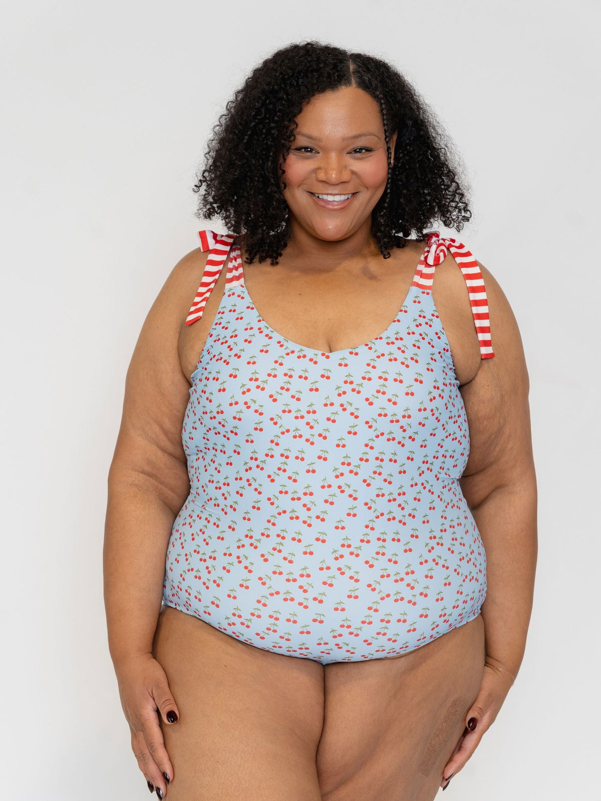 Woman wearing a light blue swimsuit with red cherry pattern and red and white striped straps on a plain background