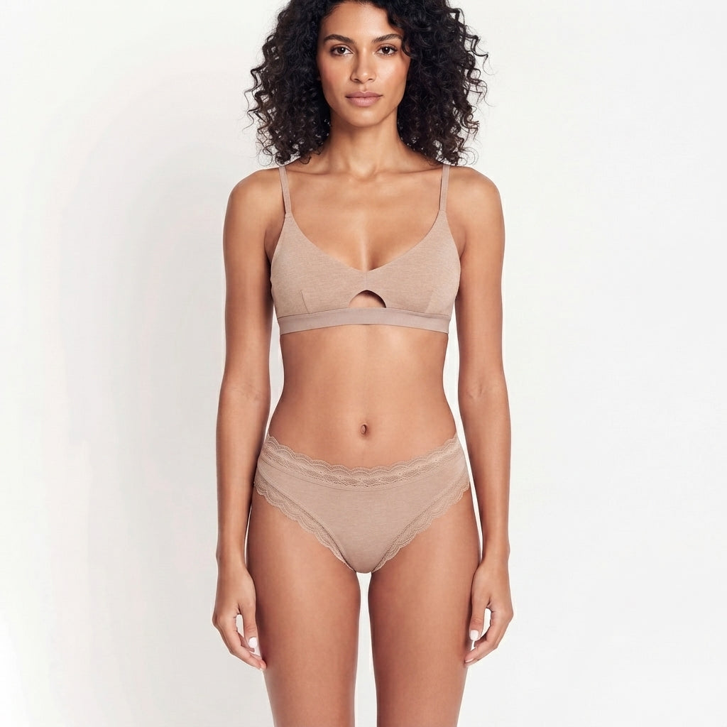 A woman modeling a matching beige lingerie set consisting of a bralette with a center cutout and high-waisted briefs, set against a plain white studio background.