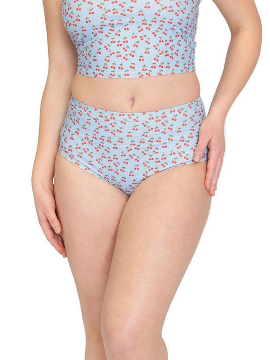 A model wearing a two-piece bikini set with a light blue background and a small red cherry print, shown from the chest down on a plain white background.