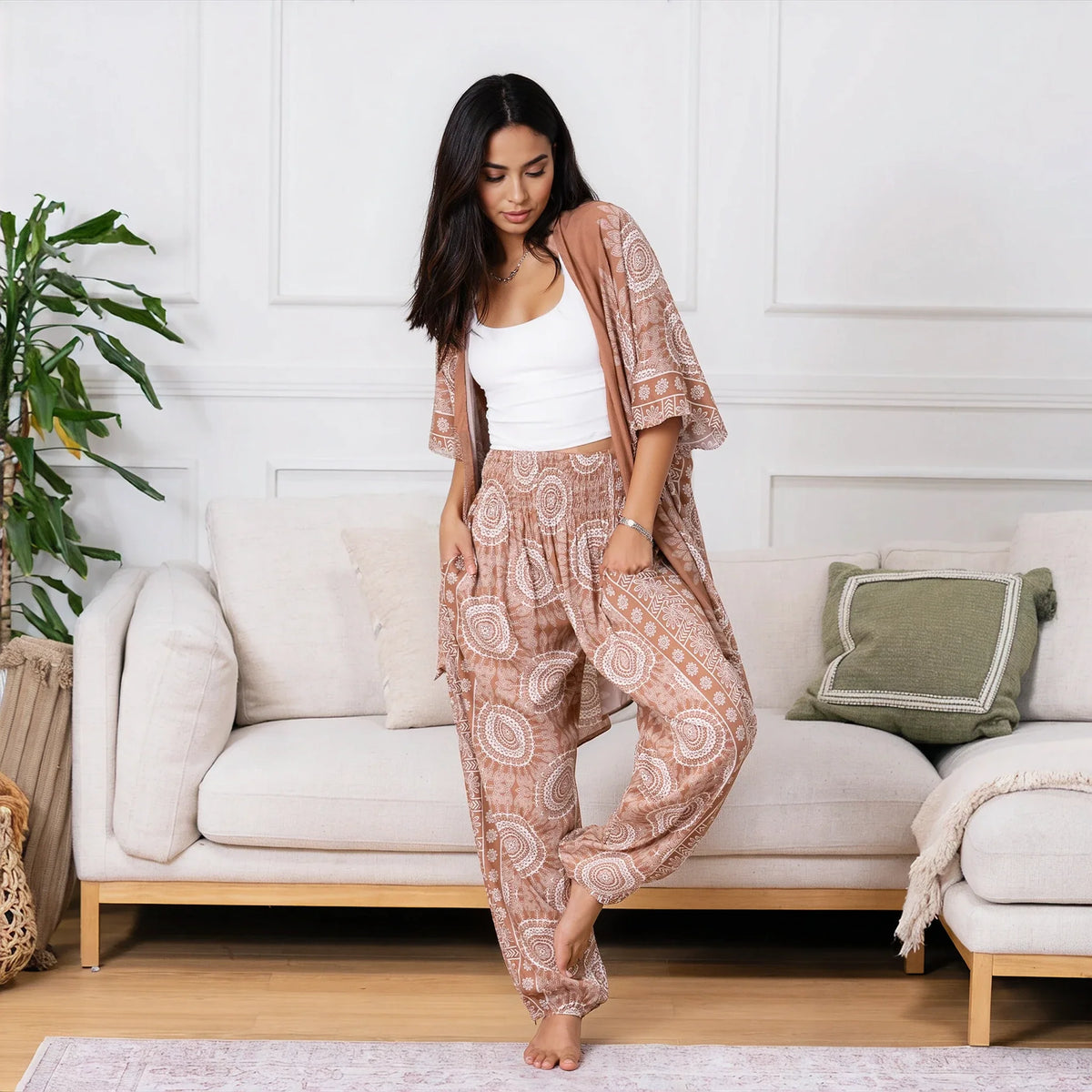 A woman stands barefoot in a bright living room wearing a matching bohemian-style lounge set consisting of a white tank top, patterned harem pants, and a matching open-front shawl. The set features a dusty rose and beige paisley pattern.