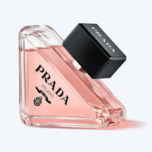 Prada Milano perfume bottle with a triangular clear glass body filled with pink liquid, a black rectangular cap, and visible PRADA branding on the bottle and cap, shown on a light gray background.