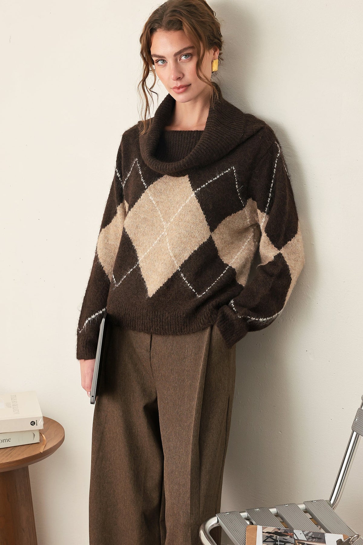 A woman wearing a dark brown cowl-neck sweater with a beige and brown argyle pattern, paired with brown wide-leg trousers, standing in a minimalist indoor setting.