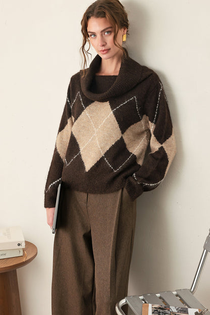 A woman wearing a dark brown cowl-neck sweater with a beige and brown argyle pattern, paired with brown wide-leg trousers, standing in a minimalist indoor setting.