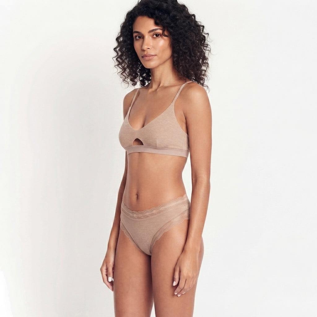 A woman modeling a matching beige lingerie set consisting of a bralette with a side cutout and high-waisted briefs, set against a plain white studio background.