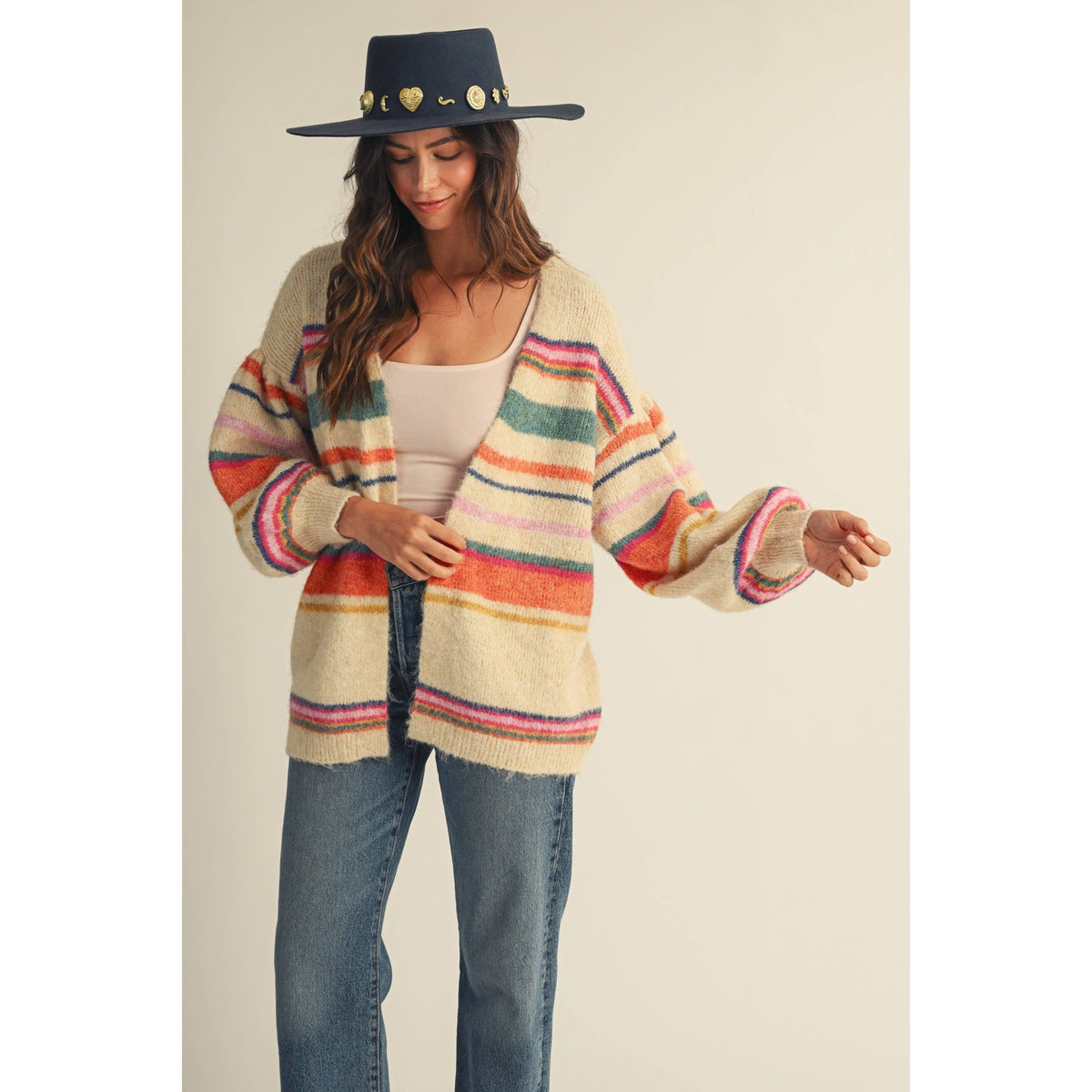 A woman wearing a cream-colored fuzzy knit cardigan with colorful horizontal stripes in pink, orange, green, and blue, paired with blue jeans and a dark navy wide-brim hat with gold heart details, set against a plain beige studio background.
