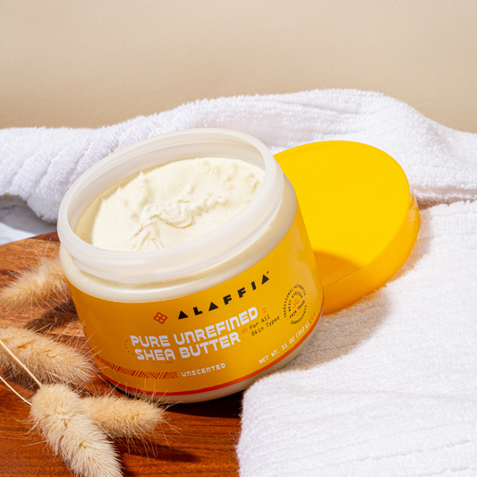 Alaffia pure unrefined shea butter jar with a yellow spatula on a wooden surface.
