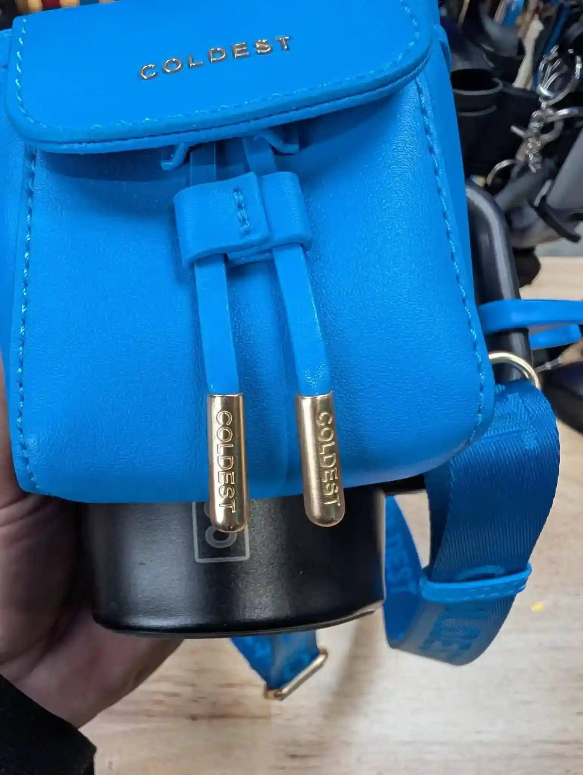 Blue backpack with 'Coldest' branding held by a hand on a wooden surface.