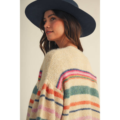 A woman wearing a cream knit sweater with colorful horizontal stripes in pink, orange, green, and blue, paired with a wide-brimmed dark navy hat, shown against a plain light beige background.