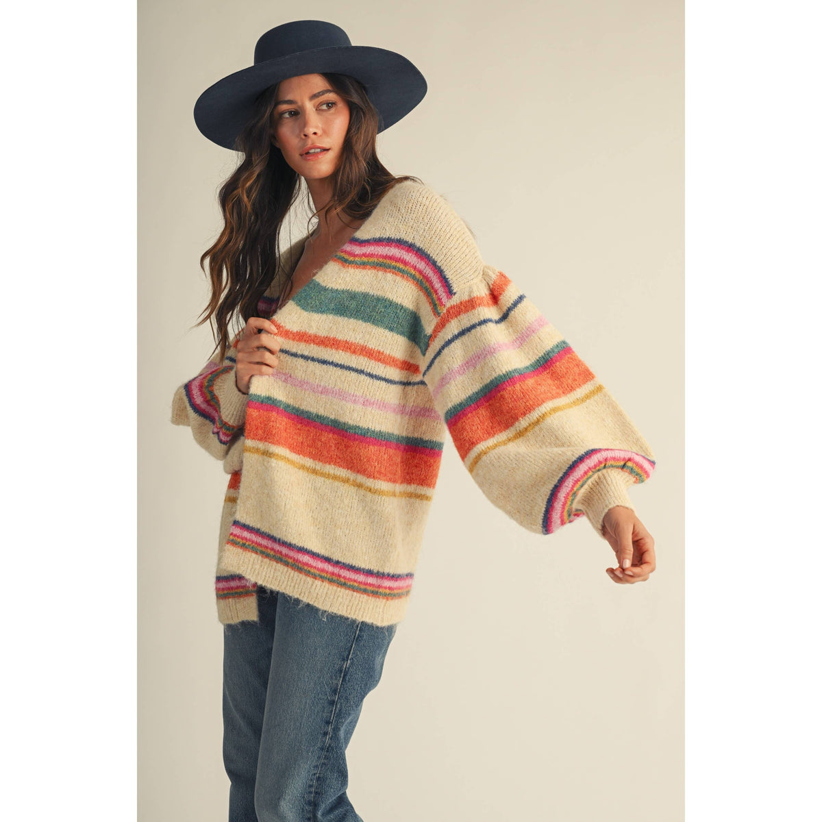 A woman wearing a cream knit sweater with horizontal multicolor stripes in pink, orange, green, and blue, paired with blue jeans and a black wide-brim hat, against a plain beige background.