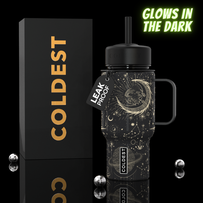 A black insulated tumbler with a handle and straw, decorated with gold celestial artwork including a crescent moon and stars. It stands beside a tall black box with large gold 'COLDEST' lettering.