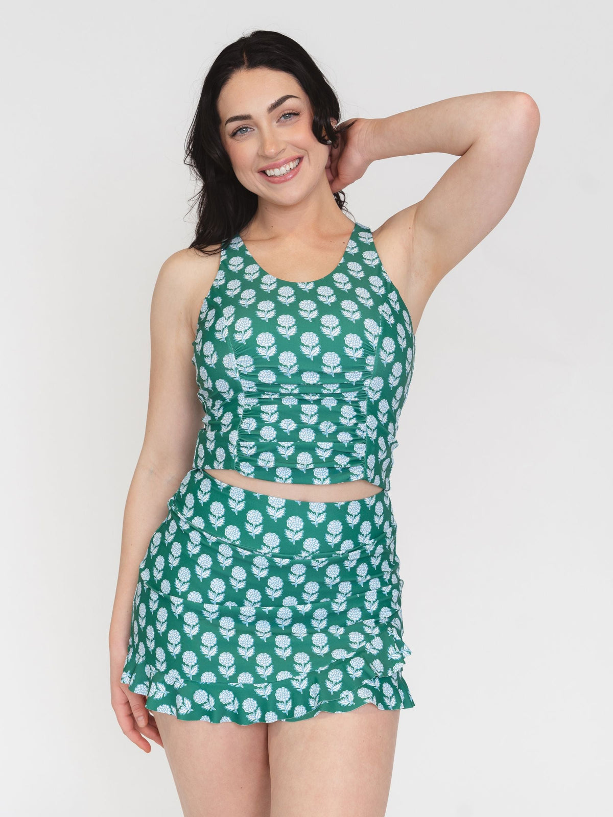 A woman modeling a matching two-piece swimsuit set featuring a green halter crop top and ruffled mini skirt, both printed with a white floral pattern, against a plain light gray background.