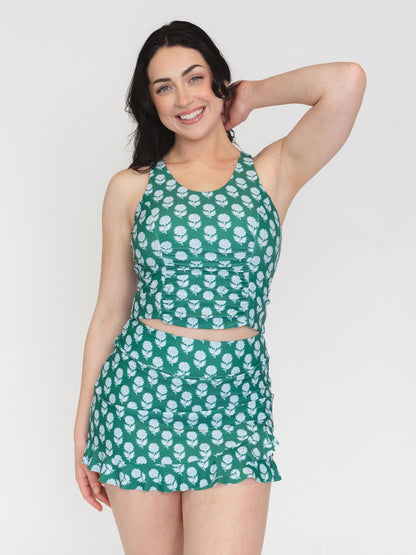 A woman modeling a matching two-piece swimsuit set featuring a green halter crop top and ruffled mini skirt, both printed with a white floral pattern, against a plain light gray background.
