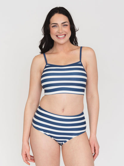 A woman modeling a two-piece striped bikini set against a plain light gray studio background. The bikini features horizontal navy blue and white stripes, with a scoop-neck crop top and high-waisted bottoms.