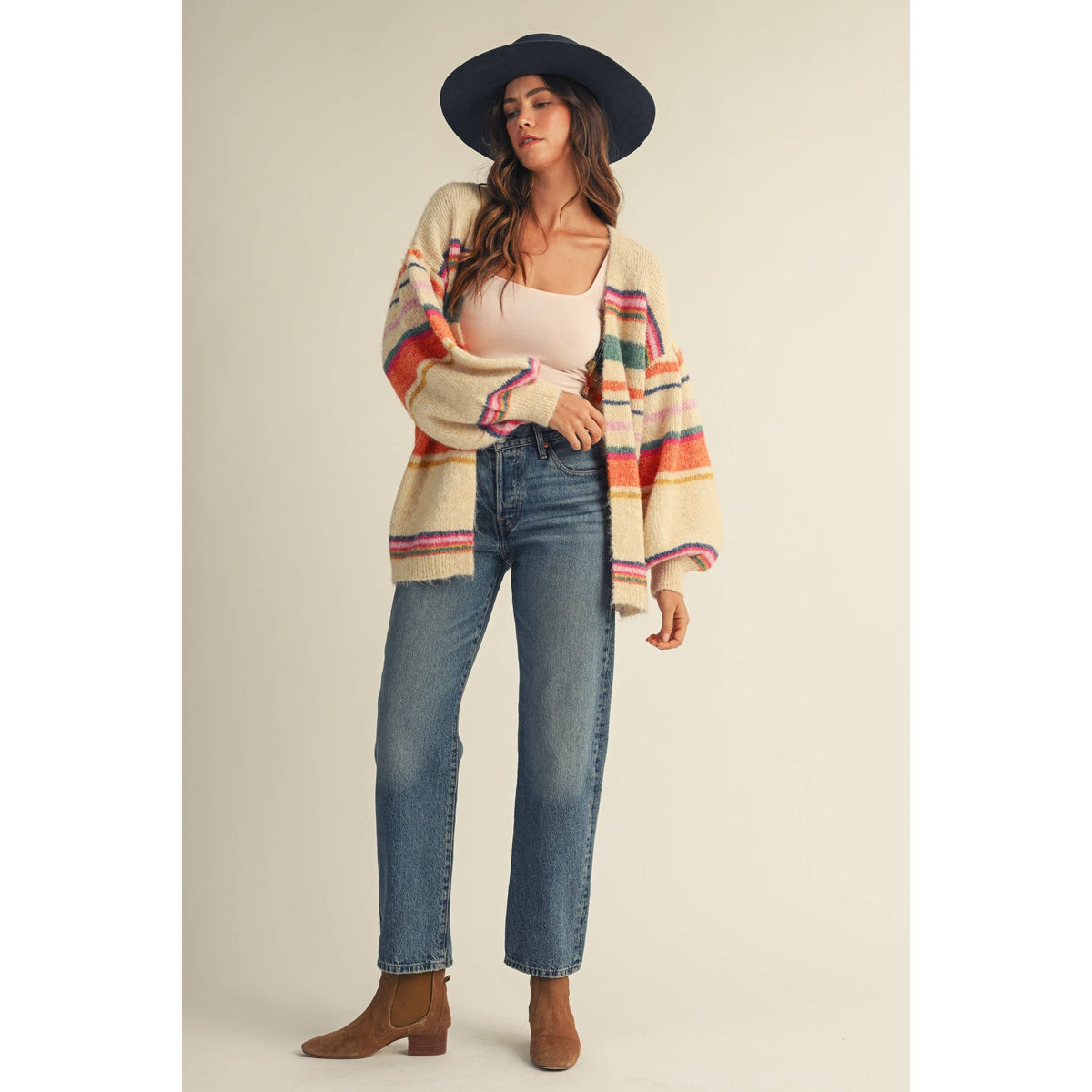 A woman modeling a cream knit cardigan with colorful horizontal stripes in orange, pink, green, and blue, paired with a black wide-brim hat, beige top, blue jeans, and brown ankle boots against a solid beige studio background.