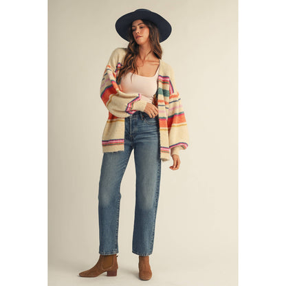 A woman modeling a cream knit cardigan with colorful horizontal stripes in orange, pink, green, and blue, paired with a black wide-brim hat, beige top, blue jeans, and brown ankle boots against a solid beige studio background.