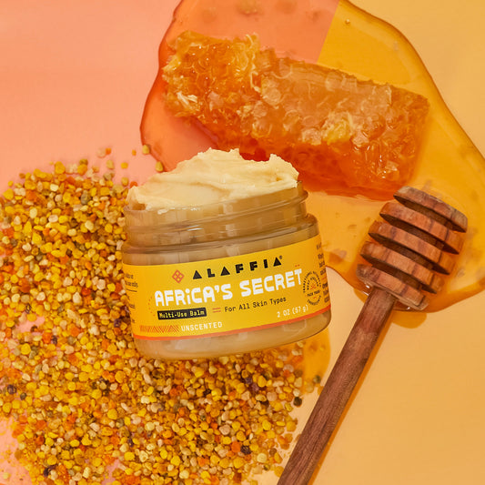 Alaffia Africa's Secret balm jar with honeycomb, honey dipper, and flowers on a peach background