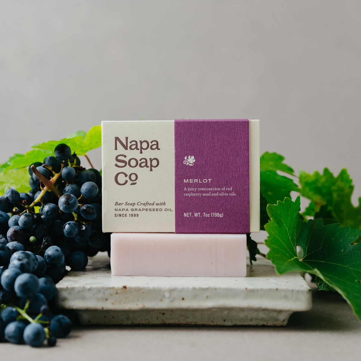 A bar of soap from Napa Soap Co. in the Merlot scent, displayed on a stone tray with grape leaves and dark grapes around it.