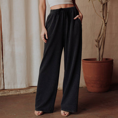 A person wearing dark charcoal wide-leg drawstring pants, standing in a rustic indoor setting with a terracotta planter and beige curtain in the background.