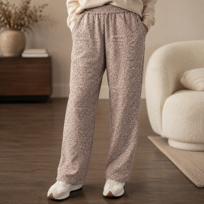 A person wearing loose, wide-leg lounge pants with a beige and taupe leopard print pattern, standing in a cozy living room setting.