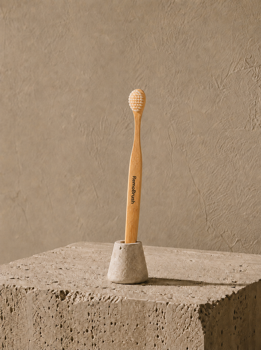 A bamboo toothbrush with a light wood handle and natural bristles stands upright in a small concrete holder on a textured stone pedestal against a warm beige plaster wall.