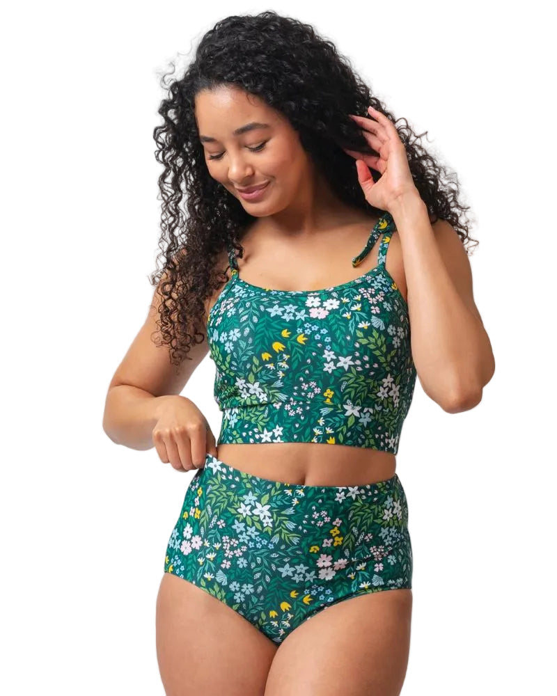 A woman modeling a two-piece swimsuit with a green floral print, featuring a cropped tank top and high-waisted bottoms, set against a plain white background.
