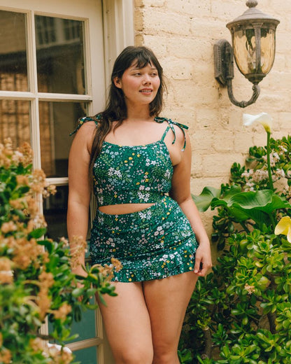 A woman modeling a green two-piece floral set consisting of a crop top and matching mini skirt, standing outdoors in front of a beige brick wall with a vintage wall lamp and green plants.