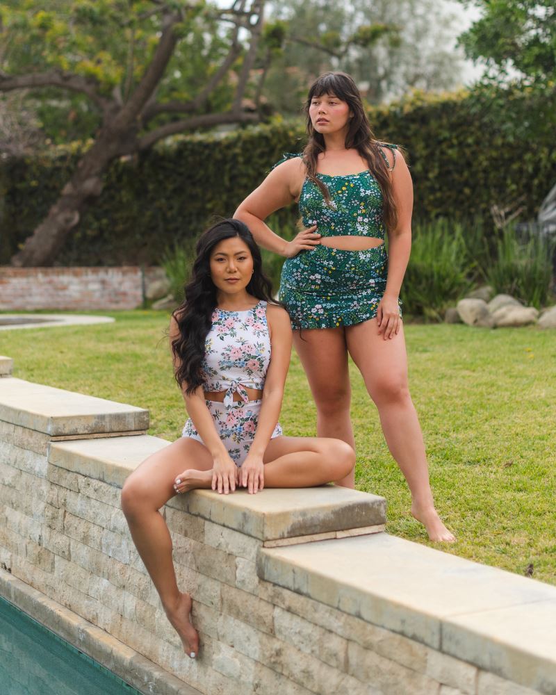 Two women in swimsuits posing by a poolside with greenery in the background