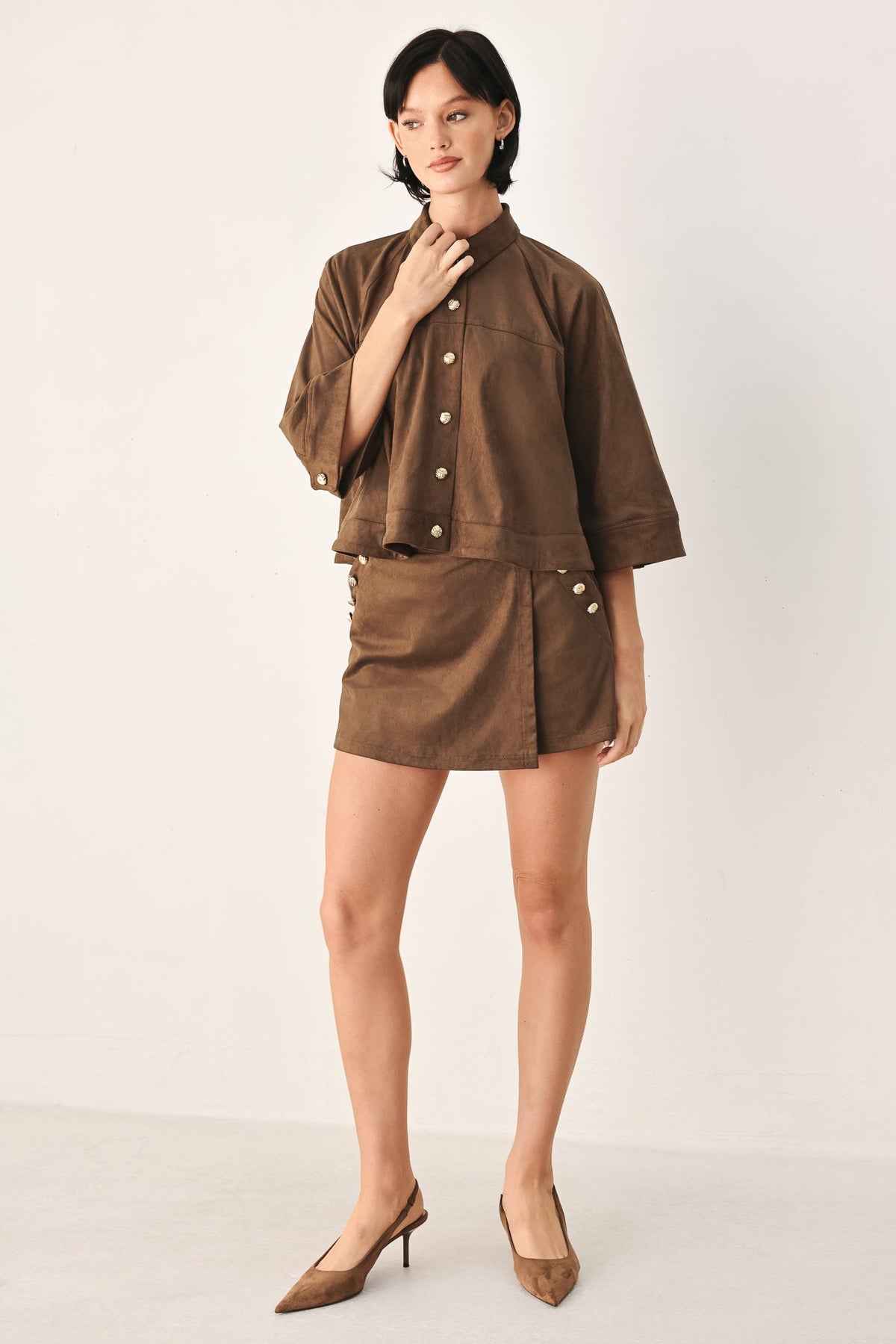 A model stands against a plain light background wearing a matching brown short-sleeve button-front jacket and high-waisted mini skirt set, paired with brown pointed-toe heels.