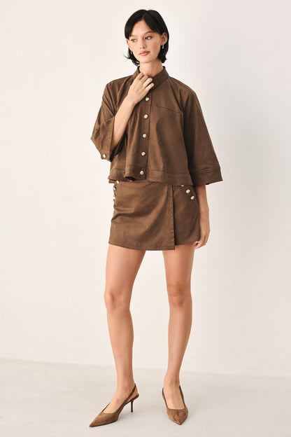 A model stands against a plain light background wearing a matching brown short-sleeve button-front jacket and high-waisted mini skirt set, paired with brown pointed-toe heels.