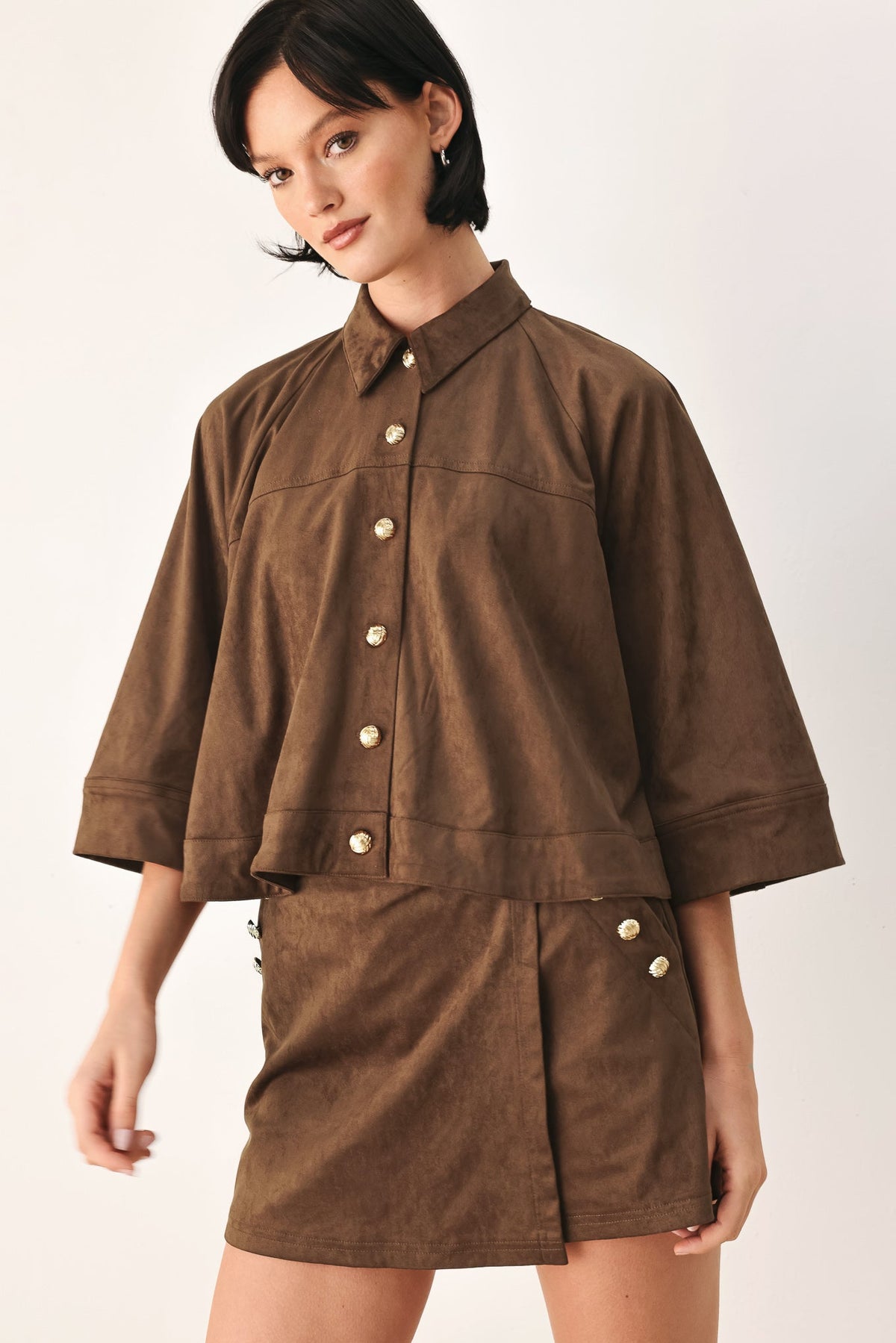 A model wearing a brown corduroy short-sleeve button-up shirt and matching brown corduroy mini skirt with decorative gold-tone buttons, set against a plain off-white studio background.
