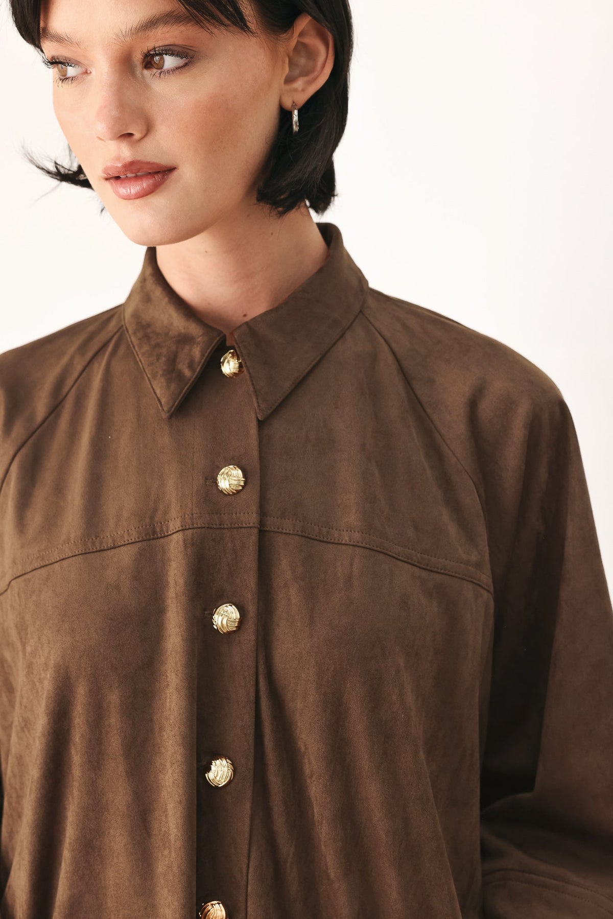 A close-up of a model wearing a brown suede button-up shirt with gold-tone buttons, set against a plain white background.