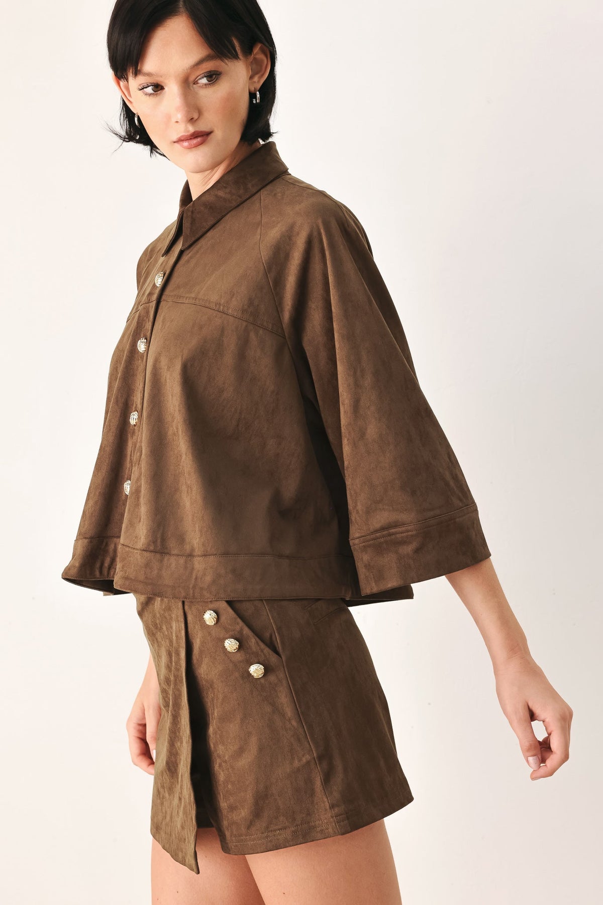 A model wearing a brown suede-like short-sleeve button-front jacket and matching shorts with decorative buttons, photographed against a plain light background.