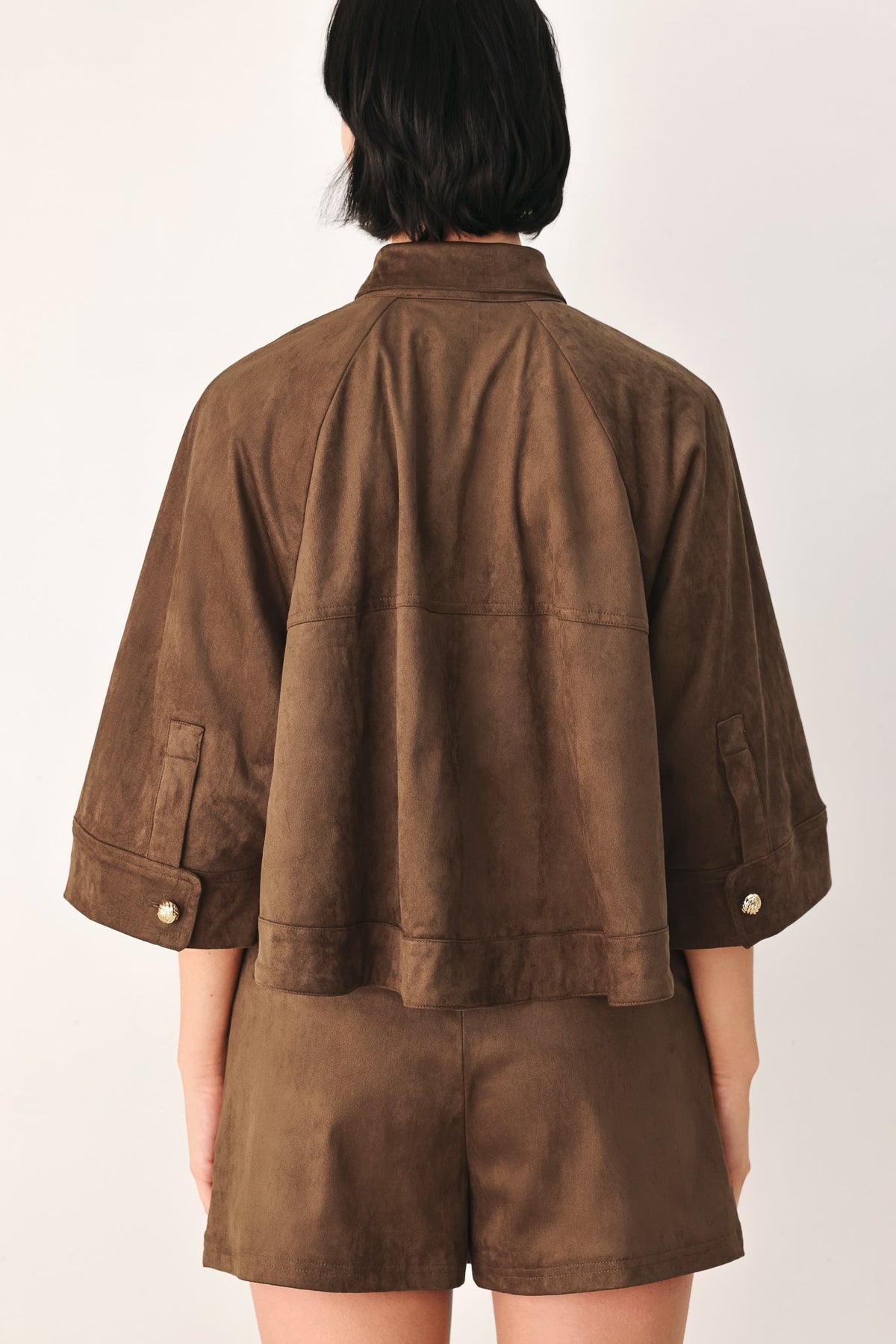 A model viewed from the back wearing a brown suede-like cropped jacket with three-quarter sleeves and matching brown shorts against a plain light background.