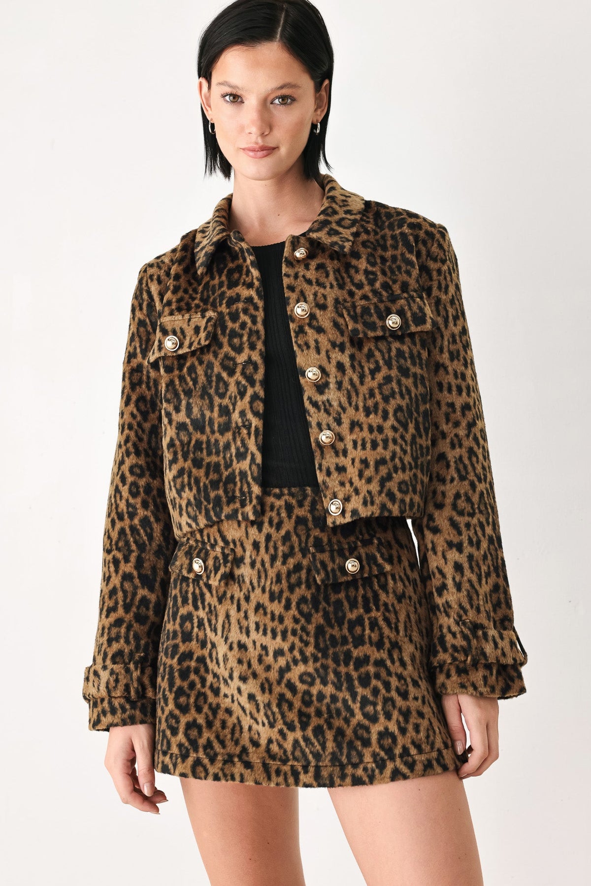 A model wearing a brown and black leopard print jacket and matching mini skirt with gold-tone buttons, styled over a black top against a plain white studio background.