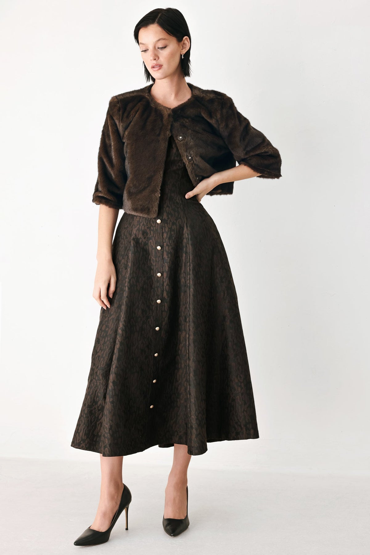 A model wearing a dark brown cropped jacket and matching dark brown button-front midi skirt, styled with black pointed-toe heels against a plain light gray studio background.