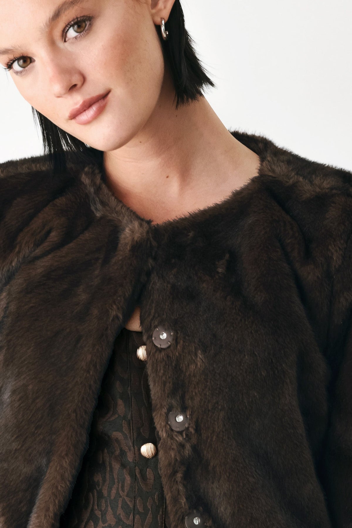 Close-up of a model wearing a dark brown fur jacket with large buttons over a patterned top, against a plain light background.