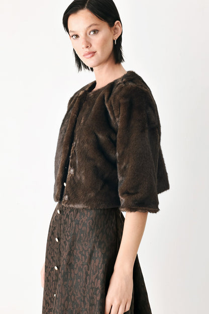 A model wearing a dark brown faux fur cropped jacket with three-quarter sleeves and a matching dark brown patterned skirt, photographed against a clean white studio background.