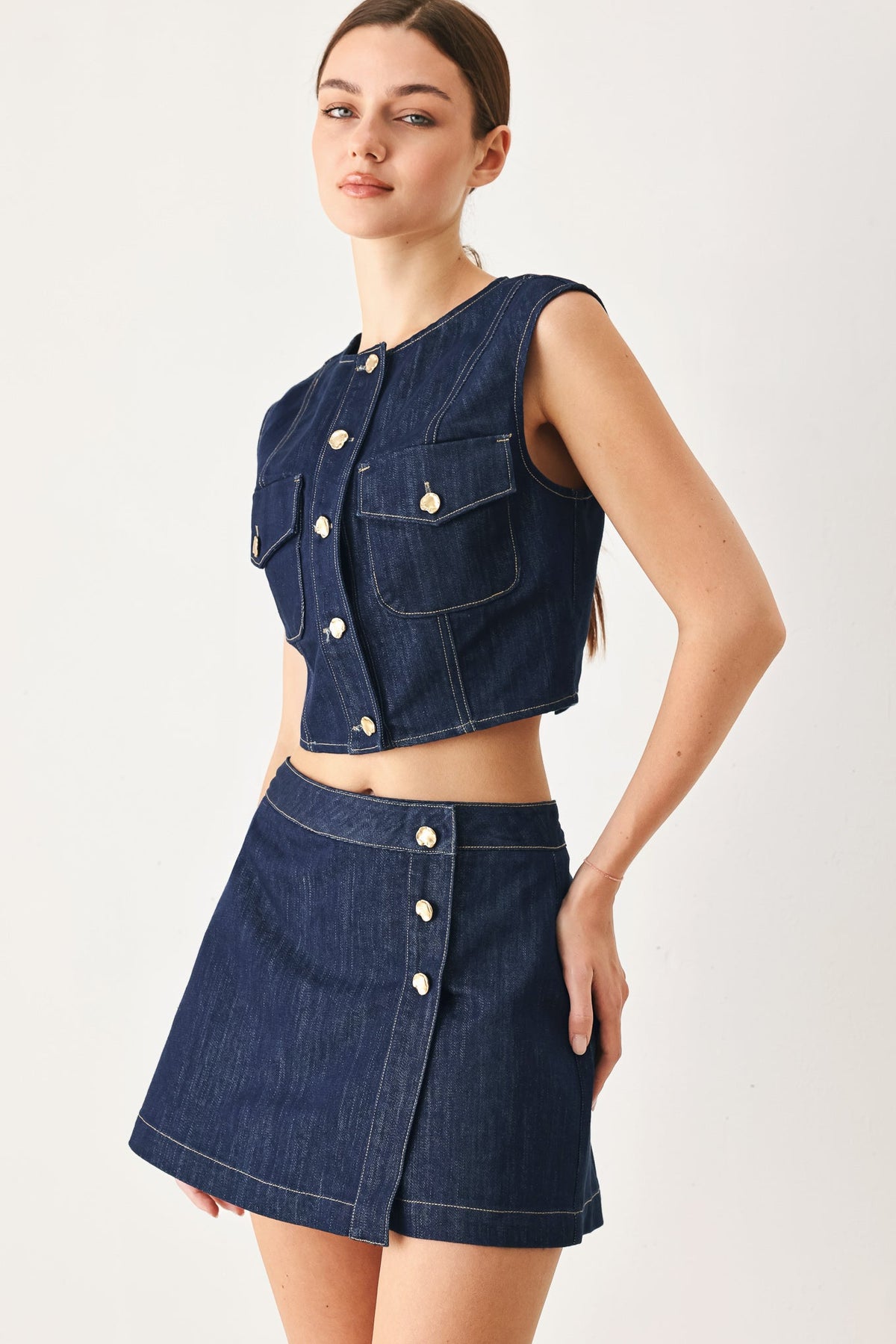 Indigo denim crop vest layered over a top, worn on a model in a light studio setting.