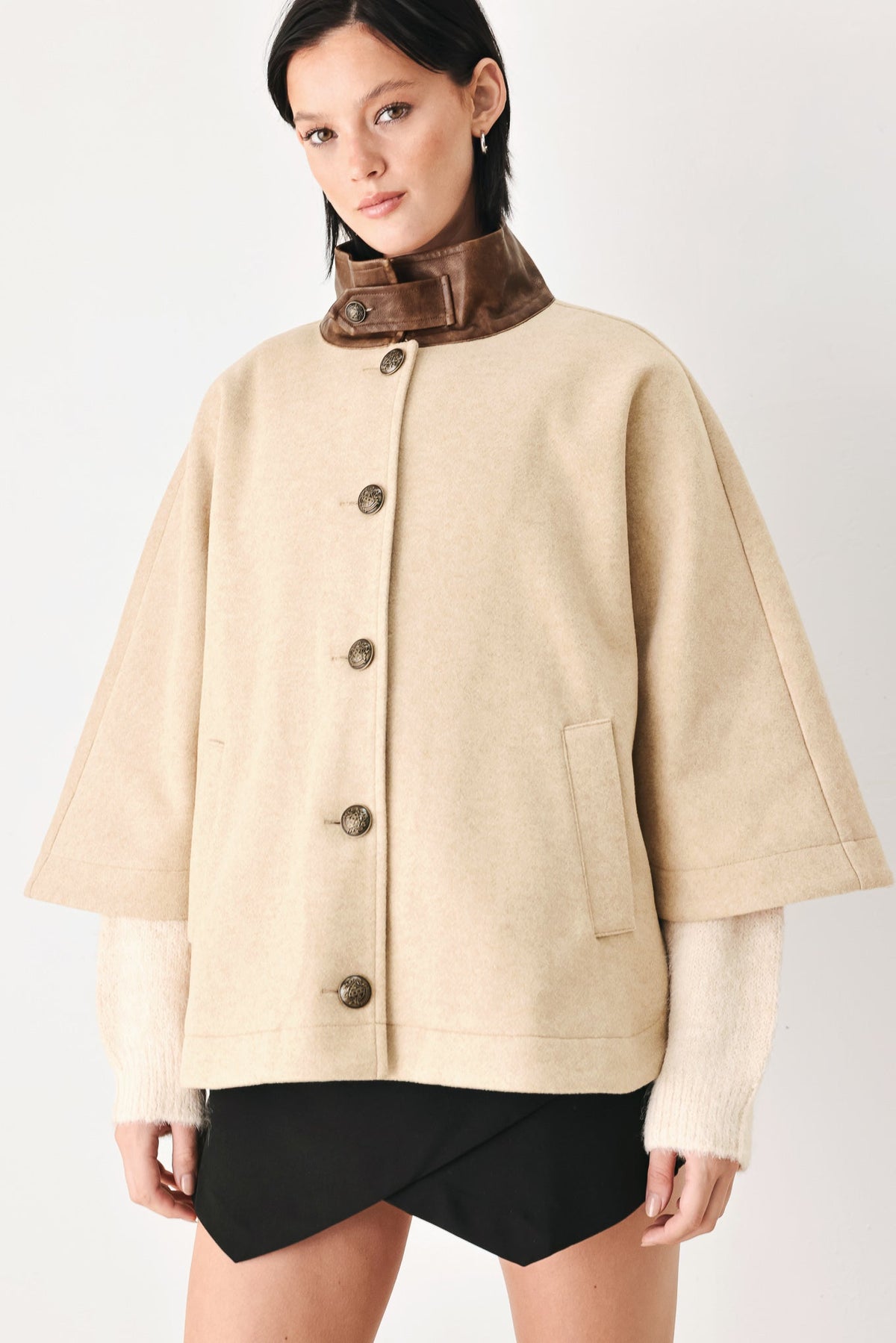 A model wearing a beige cropped cape jacket with a brown collar, buttoned front, and wide sleeves, paired with black shorts against a light gray studio background.