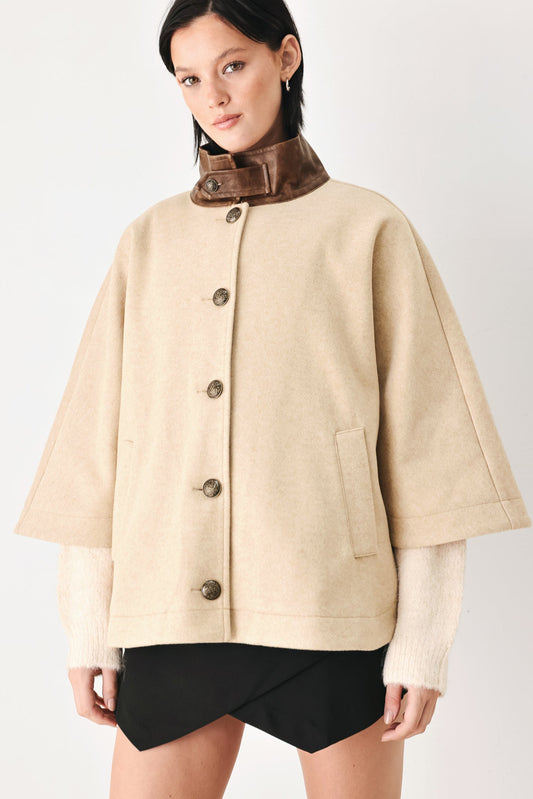 A model wearing a beige cropped cape jacket with a brown collar, buttoned front, and wide sleeves, paired with black shorts against a light gray studio background.