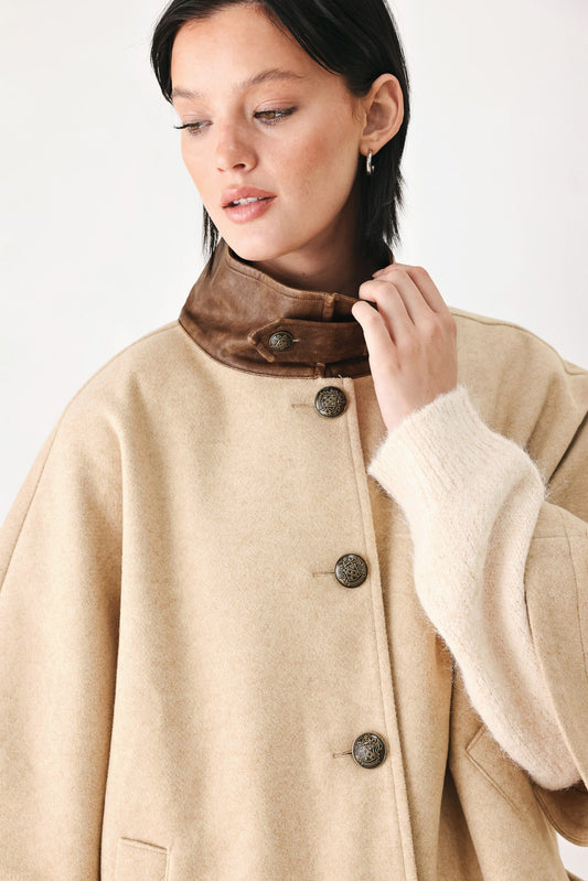 A close-up of a model wearing a beige wool coat with a high brown leather collar and dark buttons, paired with a cream knit sweater.